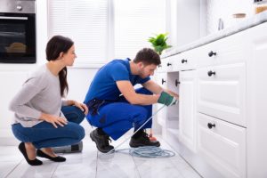Woman Looking At Male Plumber Cleaning Clogged Sink Pipe