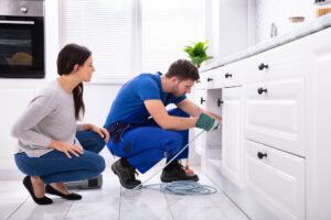 Woman Looking At Male Plumber Cleaning Clogged Sink Pipe With Drained Cable In Kitchen
