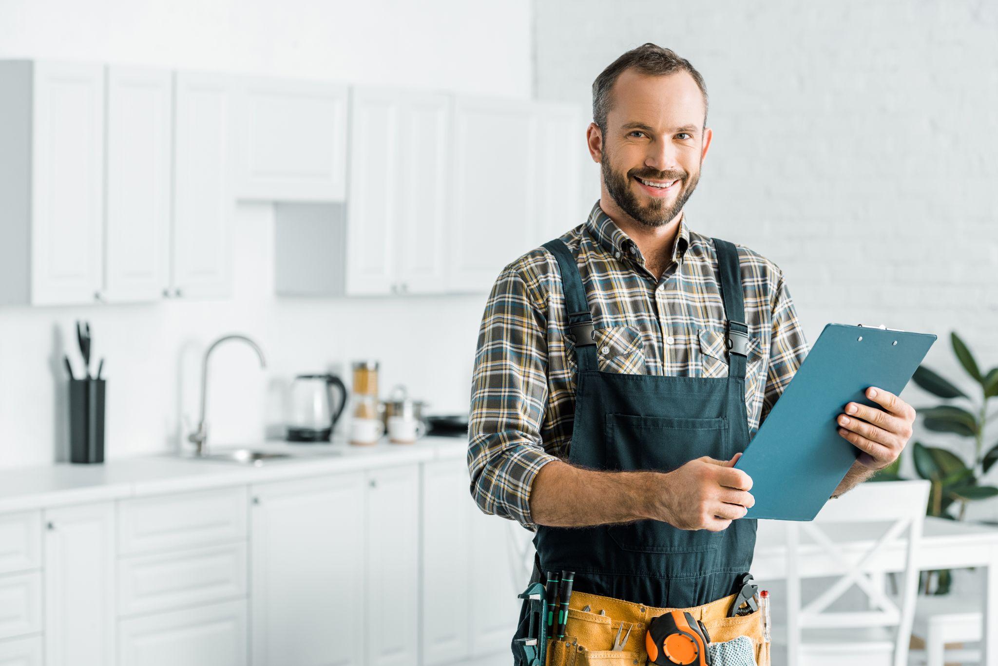 smiling plumber holding clipboard and looking at camera in kitchen