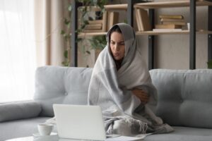 Sick ill young woman feel cold covered with blanket sit on sofa watching movie on laptop