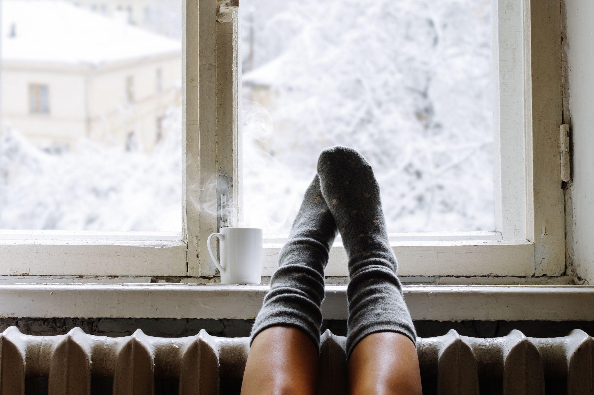 woman legs in warm woolen socks and mug of hot beverage on old windowsill against snow landscape from outside.