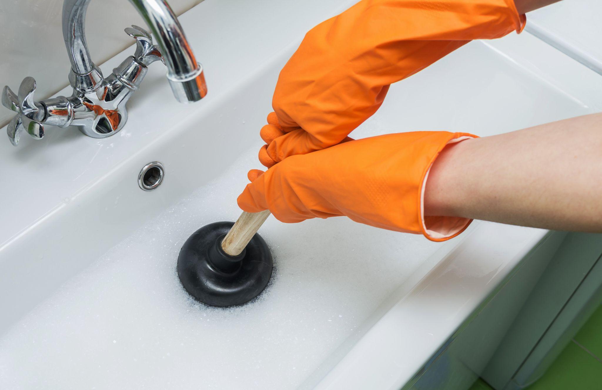 Person in protective orange gloves unblocking a clogged sink with plunger or rubber pump.
