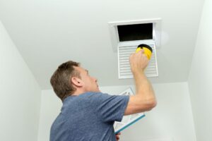 Man inspecting air ducts shining a flashlight through a small square ceiling vent into ducting pipes.